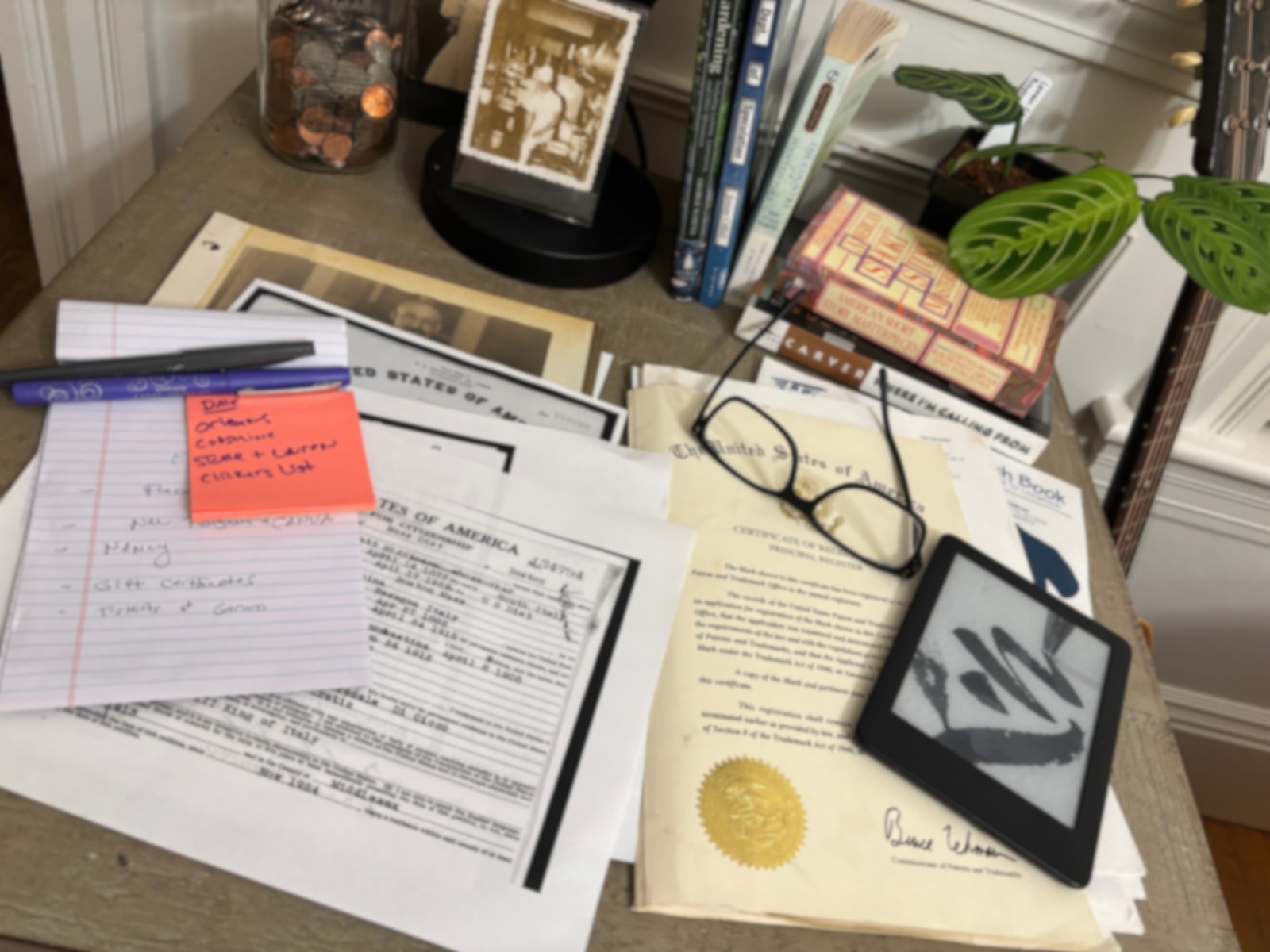 A working table covered with handwritten notes, books, historical documents, reading glasses, a Kindle, and plant research.